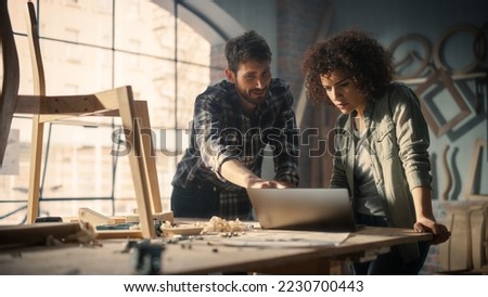 Two Young Small Business Owners Using Laptop Computer and Discussing the Design of a New Wooden Chair in a Furniture Workshop. Carpenter and a Young Female Apprentice Working in Loft Studio.