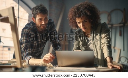 Two Young Small Business Owners Using Laptop Computer and Discussing the Design of a New Wooden Chair in a Furniture Workshop. Carpenter and a Young Female Apprentice Working in Loft Studio.
