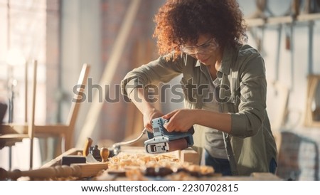 Female Carpenter Wearing Protective Safety Glasses and Using Electric Belt Sander to Work on a Wood Bar. Artist or Furniture Designer Working on a Product Idea in a Workshop. Royalty-Free Stock Photo #2230702815