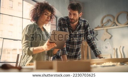 Two Talented Small Business Owners Using Tablet Computer and Discussing the Design of a New Wooden Chair in a Furniture Workshop. Carpenter and a Young Female Apprentice Working in Loft Studio.