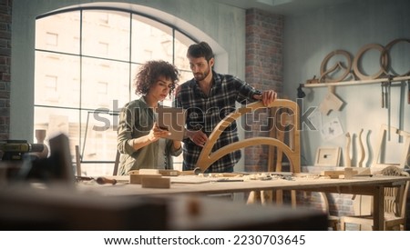 Portrait of Two Small Business Owners Using Tablet Computer and Discussing the Design of a New Wooden Chair in a Furniture Workshop. Carpenter and a Young Female Apprentice Working in Loft Studio.