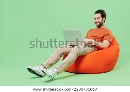 Full body young man 20s wear casual orange t-shirt sit in bag chair hold use work on laptop pc computer isolated on plain pastel light green color background studio portrait. People lifestyle concept