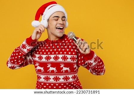 Merry fun singer young man wear red knitted christmas sweater Santa hat headphones posing listen music sing song in microphone isolated on plain yellow background. Happy New Year 2023 holiday concept Royalty-Free Stock Photo #2230731891