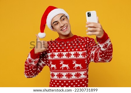 Merry young man wear red christmas sweater Santa hat posing doing selfie shot on mobile cell phone show thumb up isolated on plain yellow background. Happy New Year 2023 celebration holiday concept