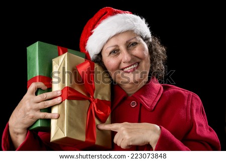 Senior woman with a smile is pointing at two golden and green wrapped Christmas presents that she is pressing against her right shoulder. Santa Claus cap and red coat. White teeth. Isolated on black. 