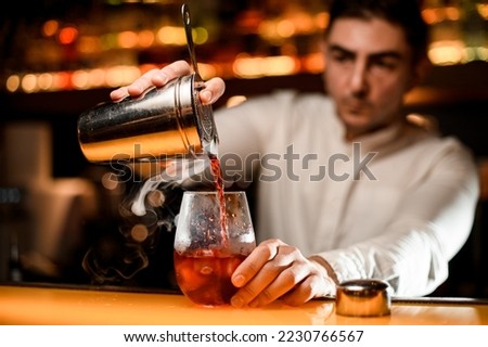 Close-up view of hand of bartender holds steel cup of shaker with strainer and pours steaming drink into glass with ice.