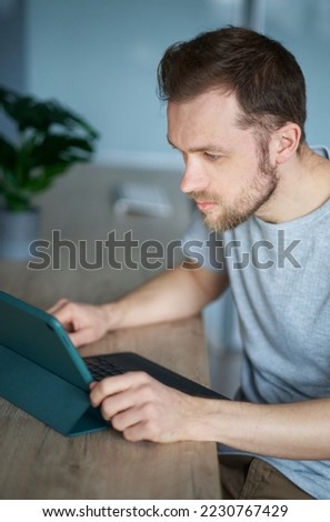 Cheerful bearded freelancer man sitting in home office using tablet working or learning online. Close-up of portrait of young handsome IT guy or engineer in grey shirt. High quality vertical photo