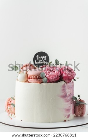Birthday cake with white cream cheese frosting decorated with red roses and pink macaroons on the white background. Happy birthday topper on anniversary cake