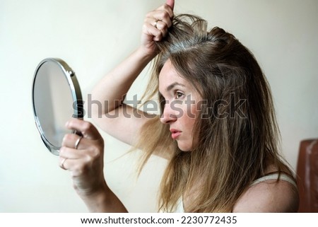 Young woman looks in the mirror at her gray hair roots. Problem of gray hair and loss hair Royalty-Free Stock Photo #2230772435