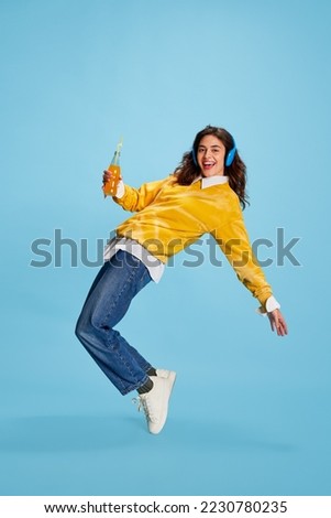 Portrait of young happy smiling girl posing in headphones isolated over blue background. Drinking juice and dancing. Concept of youth, beauty, fashion, lifestyle, emotions, facial expression. Ad