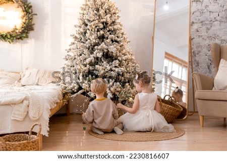 smart boy and girl play near the New Year tree, Christmas interior, open gifts, morning, lights, postcard, beige, gold, modern Scandinavian style, space for text, brother and sister, toys, wreath