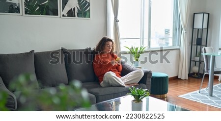 Young woman using mobile cell phone sitting on couch at home. Pretty relaxed lady holding smartphone buying in ecommerce shop, watching videos online, texting or checking social media news. Royalty-Free Stock Photo #2230822525