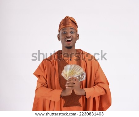 A handsome black man in his late 20s isolated on a white background African man wearing Yoruba Nigerian native traditional attire agbada excited surprised smiling happy holding money naira