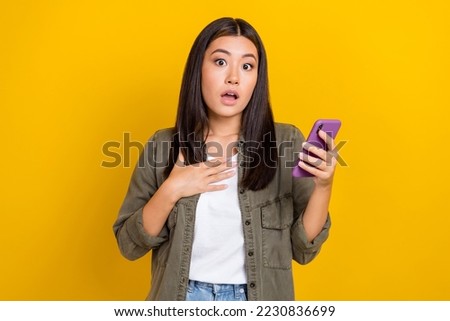 Photo of speechless impressed woman brunette hair wear gray shirt hold phone open mouth hand on chest isolated on yellow color background