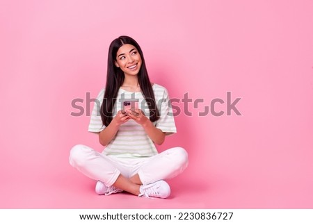 Full size portrait of cheerful pretty malaysian girl sit floor hold telephone look empty space isolated on pink color background