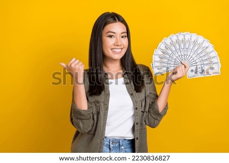 Photo of young smiling chinese girl hold money wear trendy khaki shirt finger direct mockup lottery promo isolated on yellow color background