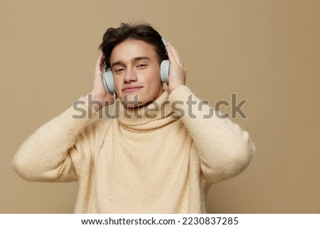 A handsome man with light white skin, with large headphones on his head with dark, short, short hair combed back, in a beige turtleneck with a high collar stands on a dark beige background.Close-up