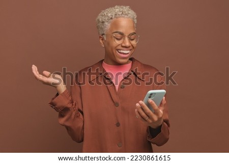 Young attractive happy African American woman joyfully looks at smartphone screen making video call via messenger or recording video message to blog subscribers stands on plain brown background