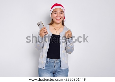 Smiling young Asian woman in a Christmas hat holding a mobile phone and doing winner gesture isolated over white background
