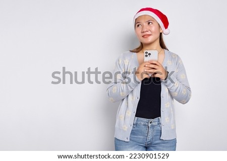 Smiling young Asian woman in a Christmas hat holding mobile phone and looking aside at copy space isolated over white background          