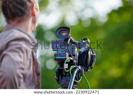 Rear view to man videographer recording outdoor movie about nature, back view to TV channel male crew with TV camera, green background. Long haired man with professional camera records video for TV Royalty-Free Stock Photo #2230922471