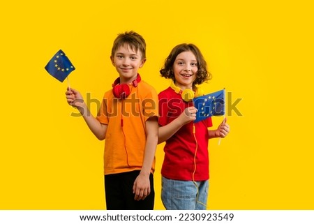 Two School Children with Headphones Hold European Union Flags. Concept Open Borders and Travel. Education excursions. Brother and Sister 9-11 y.o in Basic T-shirts Posing on Yellow Background. Royalty-Free Stock Photo #2230923549