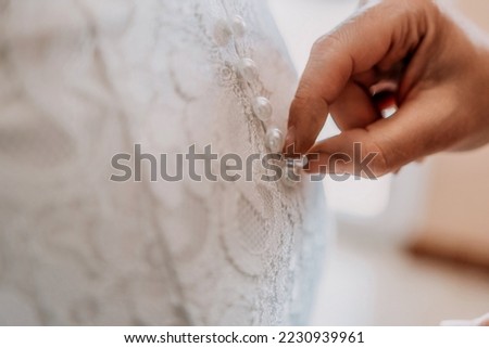 Morning preparation of the bride. Close-up of a bridesmaid, fastening a lot of buttons on the bride's wedding dress. The bride in a white wedding dress with lace standing in the room. Selective focus Royalty-Free Stock Photo #2230939961