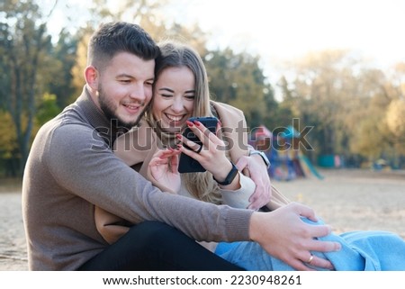 Young man and woman are sitting on the beach, looking at the phone and laughing. Horizontal photo