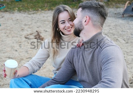 A beautiful young couple, a guy and a girl, are sitting on the beach and drinking coffee. Man and woman look at each other and smile. Horizontal photo