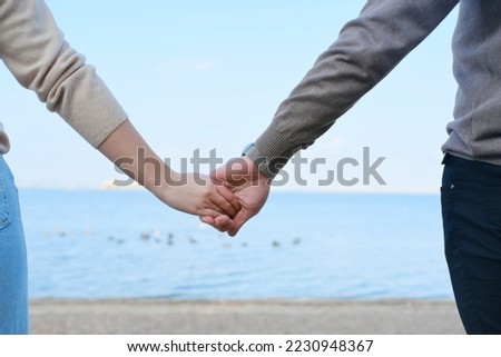 Young man and woman hold hands against the backdrop of blue water and sky. Close-up of hands. Horizontal photo