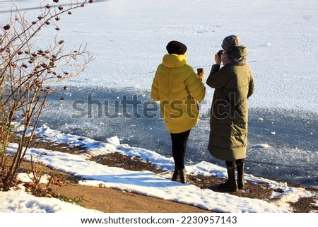 Two girls in warm coats drink hot coffee on the shore of a frozen pond, winter clothes, a country walk, a frosty winter day, keep warm with tea, a yellow jacket, friends communicate, pure snow,hats