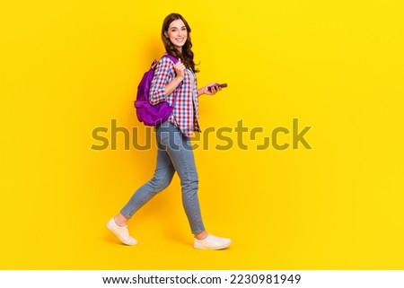 Profile full length photo of positive cheerful cute student girl walking casual style outfit hold smartphone isolated on yellow color background