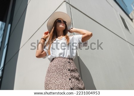 Styling beautiful young woman in fashionable summer clothes with a straw hat, sunglasses, white shirt and skirt stands near a gray building in the city in the sunlight