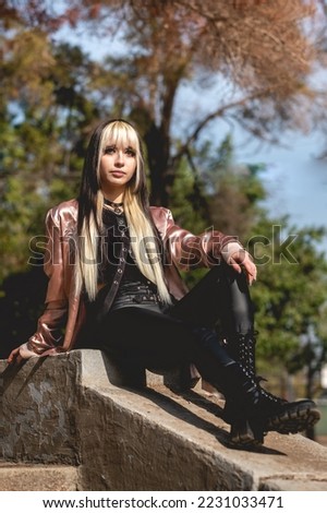 Portrait of a young and skinny model with long blonde and black hair, black clothes, pink jacket and black boots seated in the park in the day