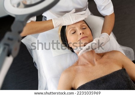 Top view of the pleasant beauty procedure with soft face massage performed by the professional therapist. Mature female laying with closed eyes 