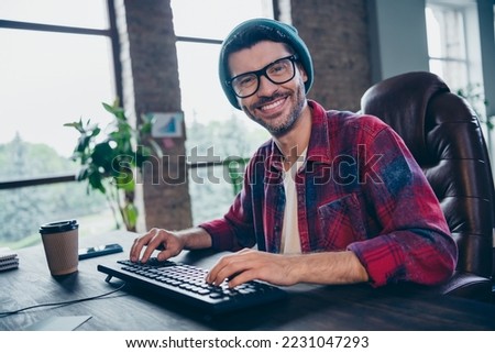 Photo of happy smiling coder dressed hat glasses typing program modern gadget indoors workplace workstation loft
