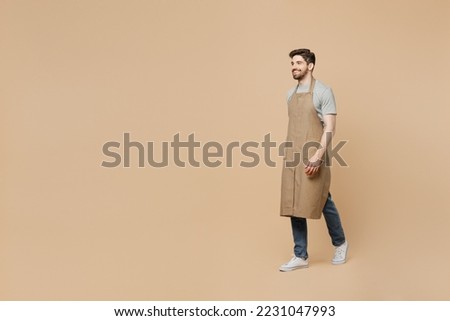 Full body side view smiling happy young man barista barman employee wear brown apron work in coffee shop walking going isolated on plain pastel light beige background. Small business startup concept