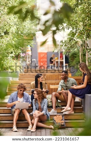 Chilling on campus between classes. A group of young students relaxing outdoors with technological devices. Royalty-Free Stock Photo #2231061657