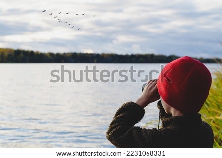 Kid on lake shore watching in binoculars how flock of migrating birds training to fly in wedge Royalty-Free Stock Photo #2231068331
