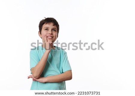Happy teenage boy wearing turquoise t-shirt, holding his hand at chin and cutely smiling, thoughtfully looking at aside, isolated over white background with copy ad space for promotional text