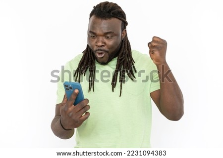Black man celebrating victory while looking at phone screen. Excited man standing against white background, raising fist, celebrating success or receiving great news. Achievement, excitement concept