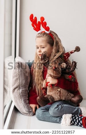 little girl with a plush deer sitting on the window. A child looks out the window and is waiting for Christmas, Santa Claus