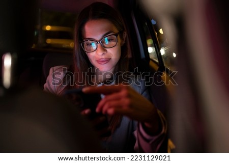 Portrait of a business woman using smartphone while sitting in a backseat of a car at night. Beautiful female passenger using mobile phone in a taxi in an urban city street with working neon signs. Royalty-Free Stock Photo #2231129021