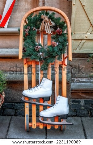 Two figure ice skates, wooden sledge, Christmas wreath . Pair of white leather skate shoes hanging, wood sled decorating with fir wreath. Vintage wintertime holidays equipment for kids, street decor