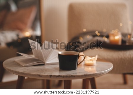 Cup of tea with paper open book and burning scented candles on marble table over cozy chair and glowing lights in bedroom closeup. Winter holiday season.  Royalty-Free Stock Photo #2231197921