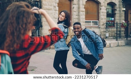 Creative girl and guy friends are posing for camera standing in the street in city while young woman with backpack is taking pictures. People are having fun and laughing.