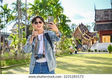 Handsome and happy young Asian male traveler or travel blogger smiling, showing thumb up, taking selfie or video with his smartphone while visiting beautiful Thai's temple.