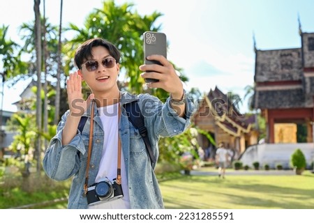 Handsome young Asian male traveler or travel blogger smiling, waving hand, taking selfie or video with his smartphone while visiting beautiful temple.