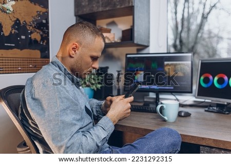 Side view of male video editor using smartphone while working on project at table with computer monitors at home 