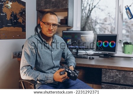 Portrait of smiling male videographer with photo camera working on video editing while sitting at table with computer monitors and looking at camera 